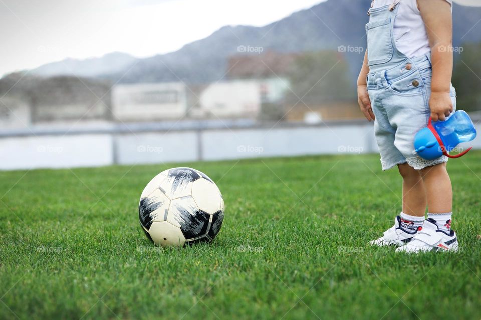 A small child with a drinking bowl in his hands on a football field and a ball.