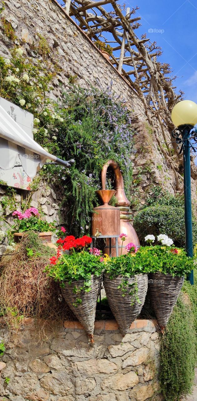 A beautiful view of a variety of flowers in wicker cone pots, a copper jug fountain and hanging plants growing on stone walls on a sunny summer day in the city of Positano, Italy, close-up side view. The concept of traditional urban plants.