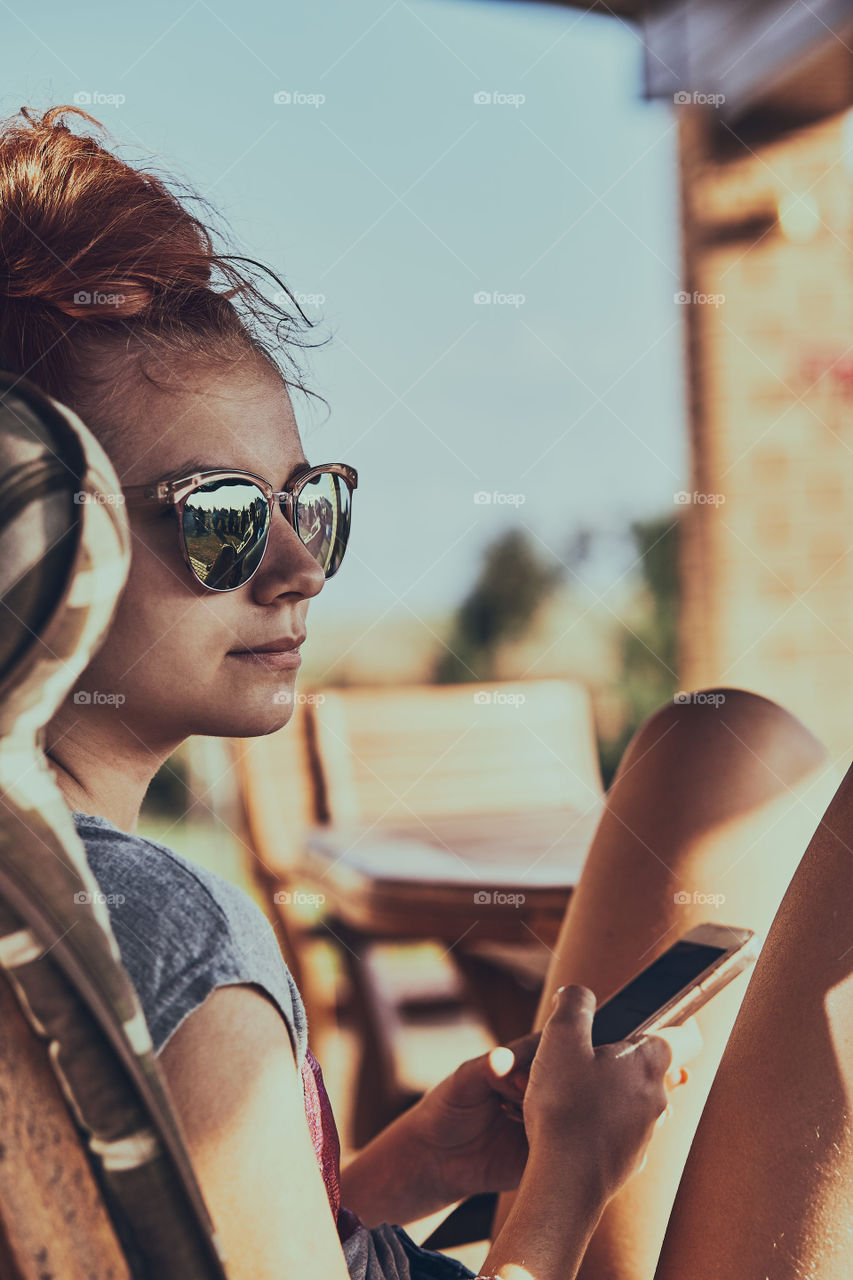 Young woman chatting on a  mobile phone sitting in a chair outdoors on patio wearing a sunglasses. Real people, authentic situations