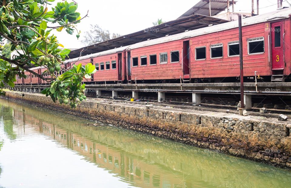 Train stop near the river. Reflection of train can be seen on the water. Row of rectangle shape box which have rectangle windows.