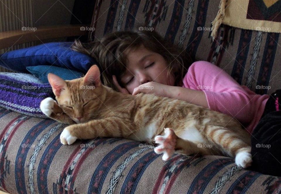 It’s nap time for all good little girls and kitties! Girl and orange tabby taking afternoon nap together on a couch