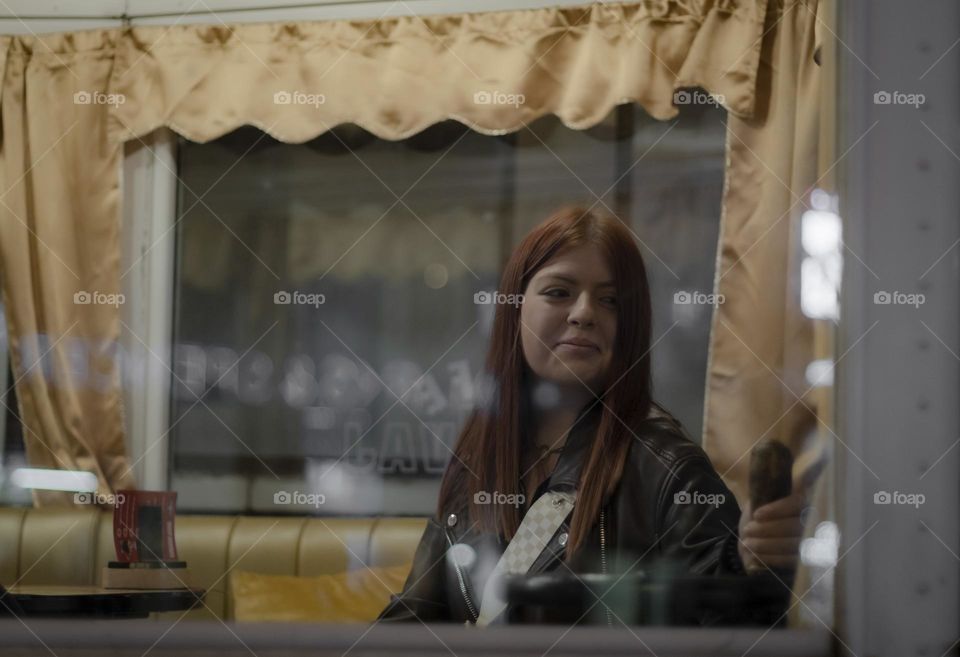 Portrait of a beautiful caucasian teenage girl with long brown hair sitting outside the window in a restaurant car and looking away with a smile at night in prague, close-up side view. The concept of people and windows.