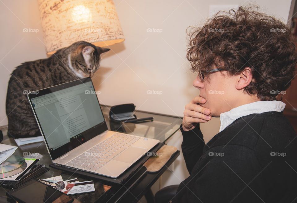 Man writing at his laptop computer pausing for a second to ponder helps next line as a cat looks on, perched on top of the desk.