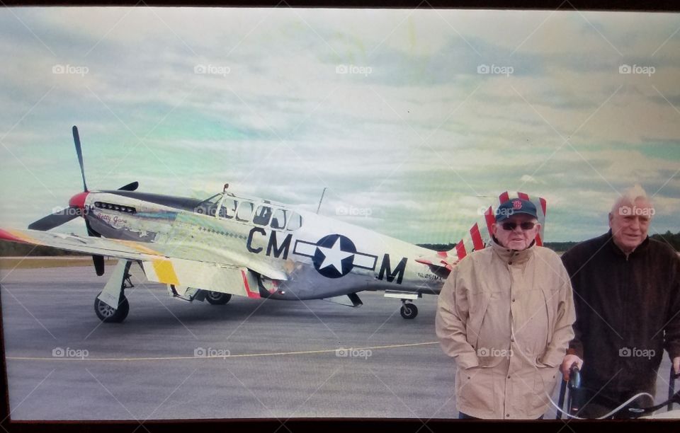Older men at airport with WWII Fighter Jet in background. Cold day, wearing coats & using walker & Oxygen tube.