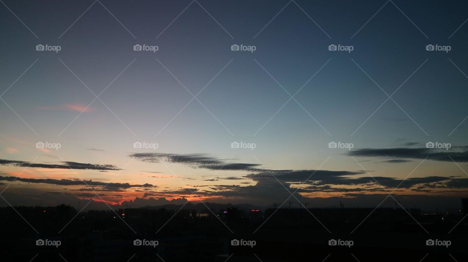 The picture shows a beautiful sunset over a city skyline. The sky is a warm orange and pink color, with clouds in the distance. The buildings in the foreground are tall and modern, with lights on in some of the windows. The overall mood of the pictur