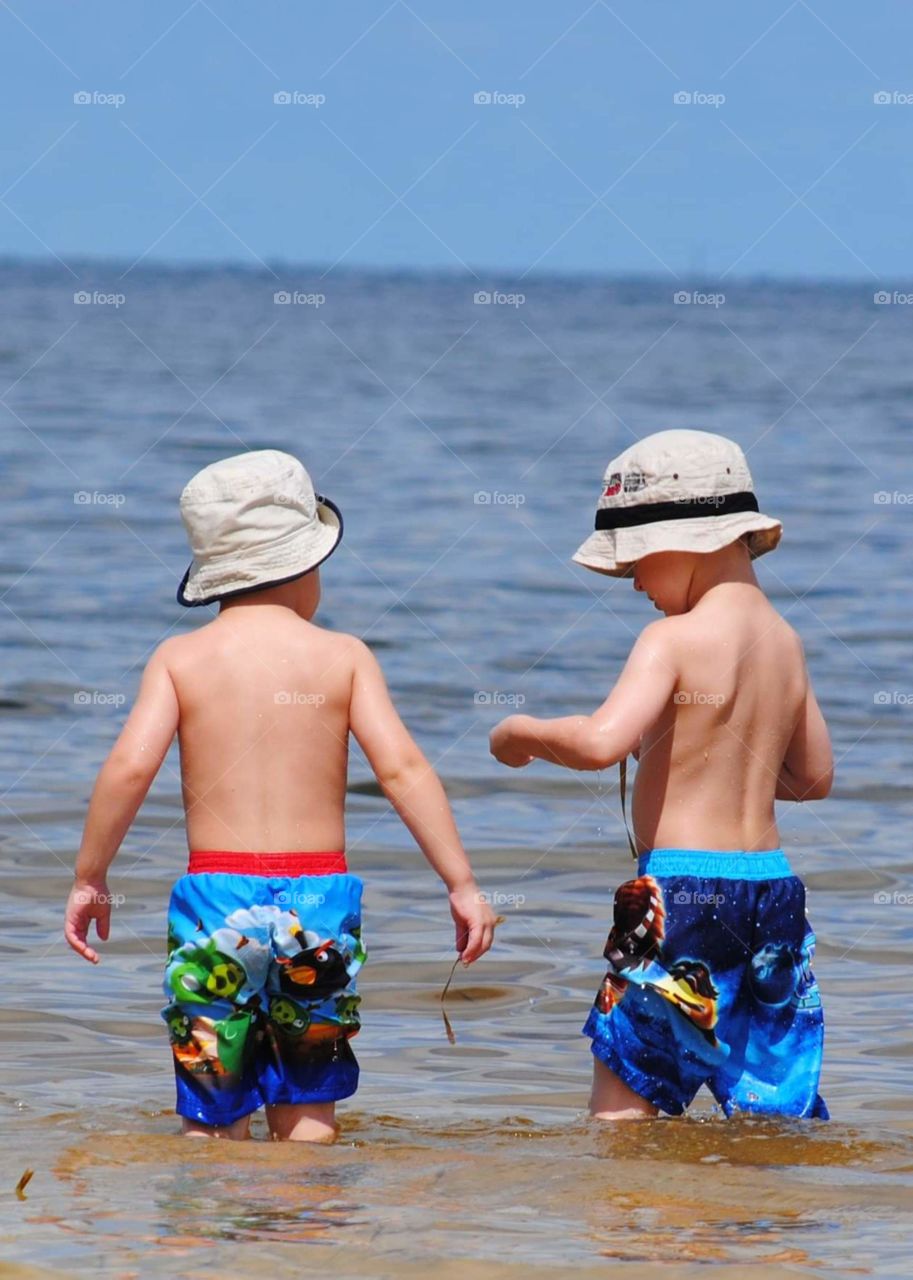 two young boys wearing blue swim shorts and tan fishing hats exploring the water's edge on a bright sunny day