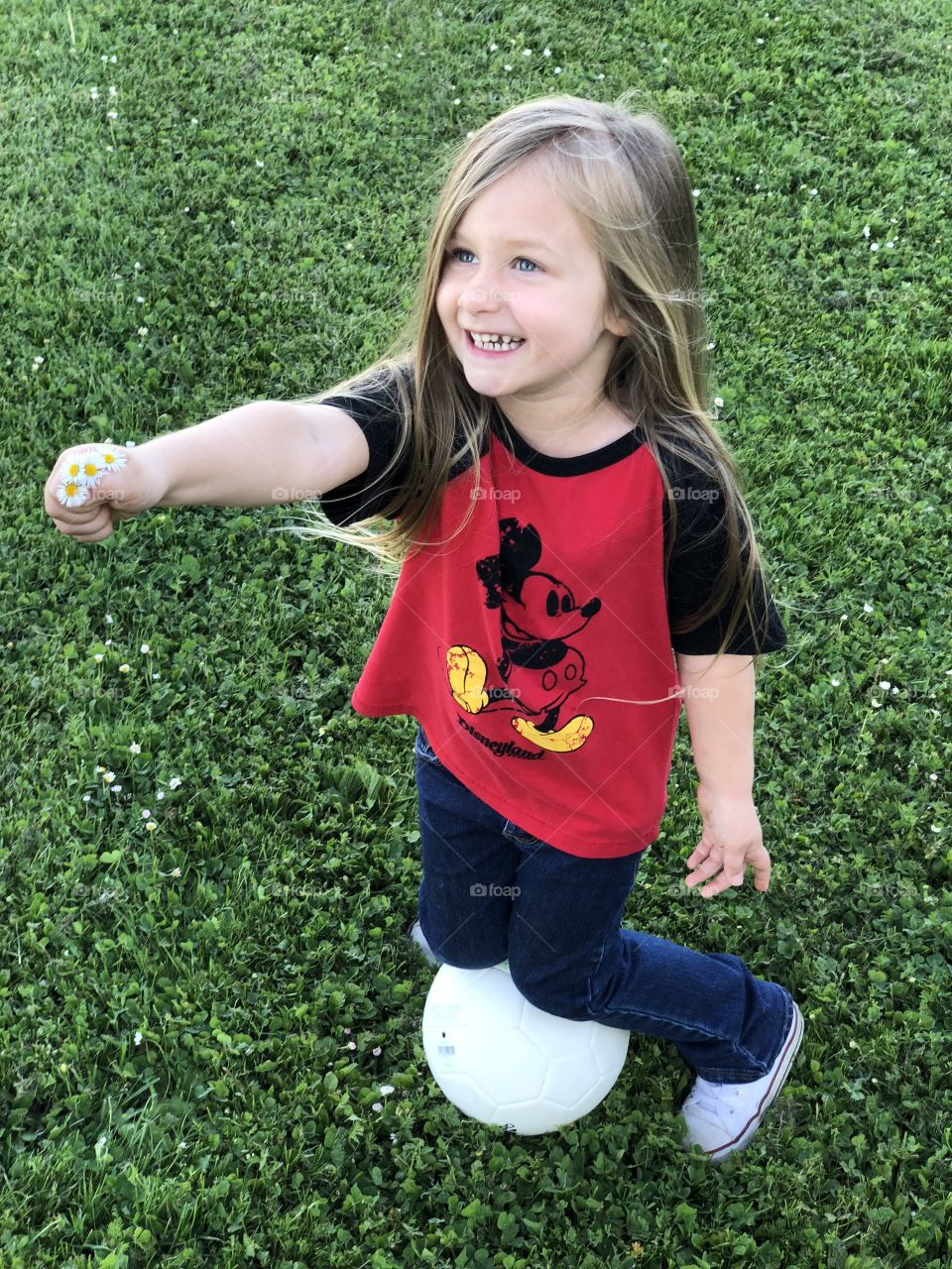 Little blue eyed girl with long blond hair stopping to show a handful of daisies kneeling on a soccer ball