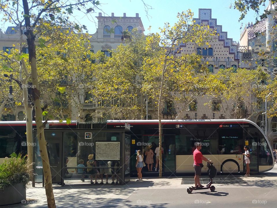 people boarding the tram as an electric scooter passes by. other people are waiting at the tram stop.