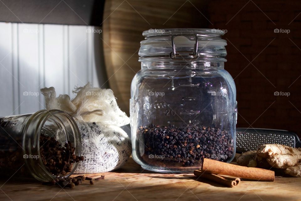 Ingredients for elderberry syrup—cloves in small glass jar, elderberries and chopped ginger in cheesecloth, dried elderberries in sealed glass jar, cinnamon sticks and ginger root with grater on wooden surface