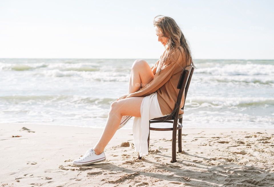 Young carefree beautiful woman with long hair in sweater sitting on chair on sea beach