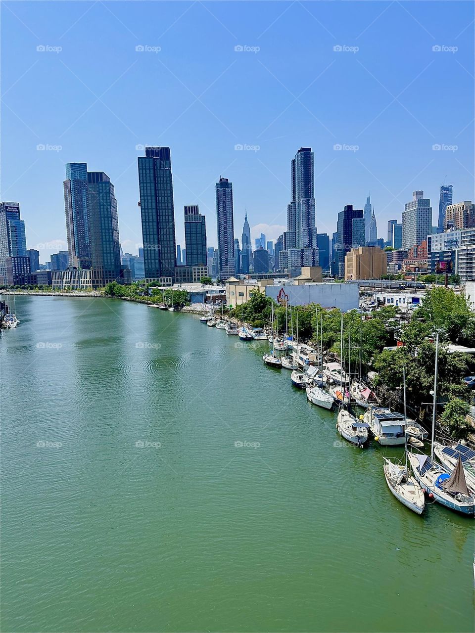 This is the “E River” and “Newtown Creek” with shore vegetation and various boats seen from the “Pulaski Bridge” that connects “Greenpoint”, Bklyn to LIC. In the distance we see “Manhattan” and the “Empire State Building”. 2024. Hypnotic Productions