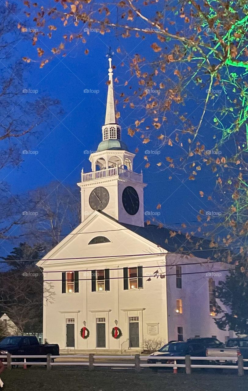 A white church glows against a deep blue post-sunset sky, accented by warm candlelit windows, twinkling Christmas lights, and a real tree outside bathed in soft green light—simple, serene, and festive.