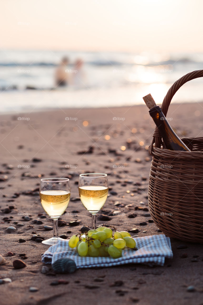 Two wine glasses with white wine standing on sand, on beach, beside grapes and wicker basket with bottle of wine. Young couple standing in the sea in the background