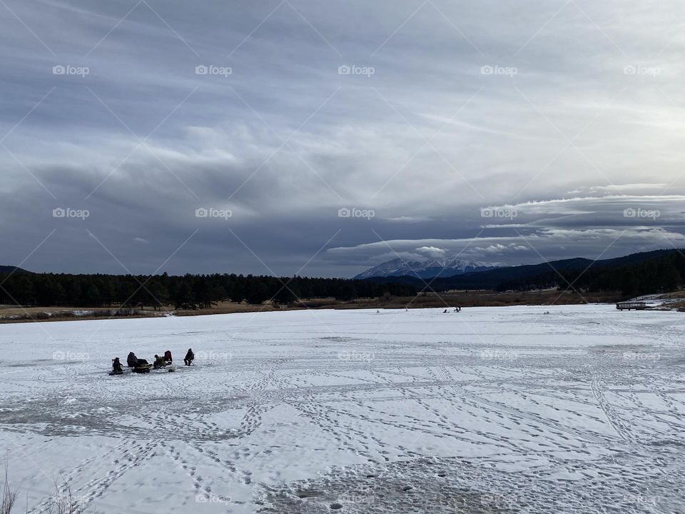 Men ice fishing on a snow covered frozen lake in the winter. Snowy Pikes peak in the background is peeking through the winter clouds. 