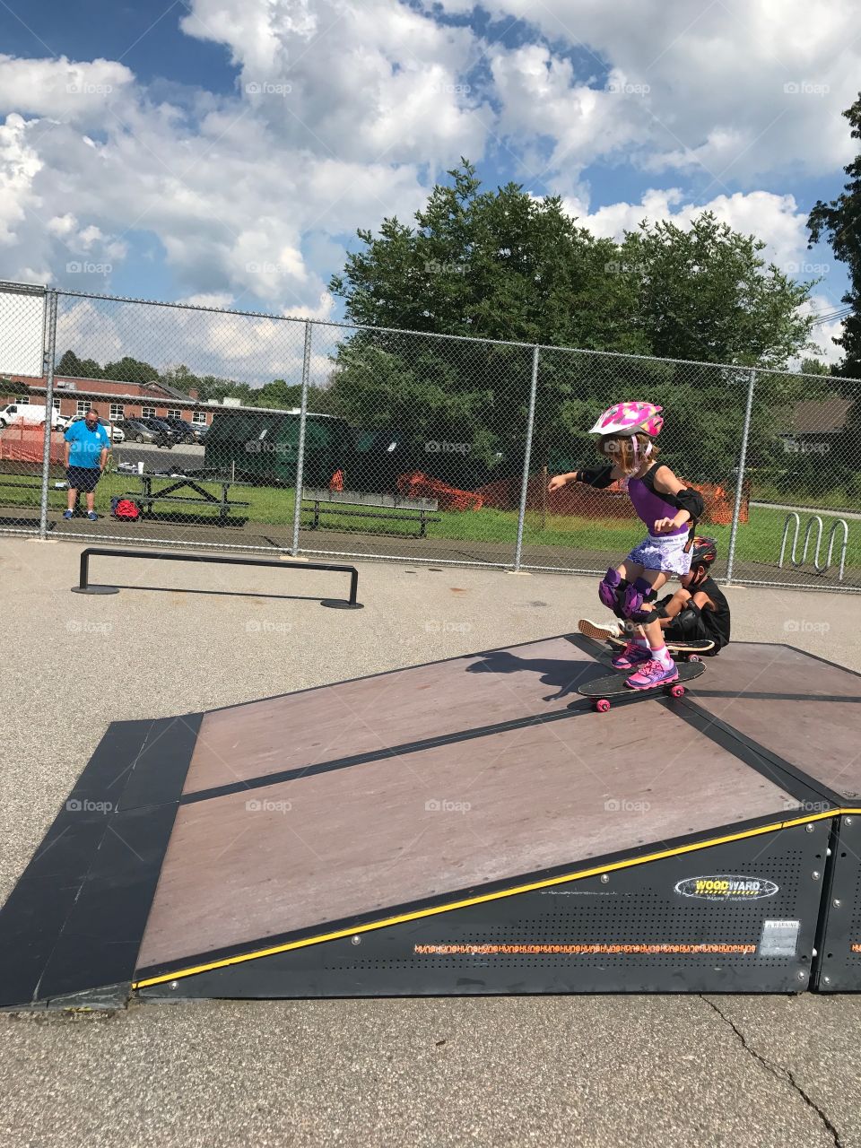 KBrave girl skateboarding on ramp in skate park with pink safety gear and brother sitting next to her to cheer her on.