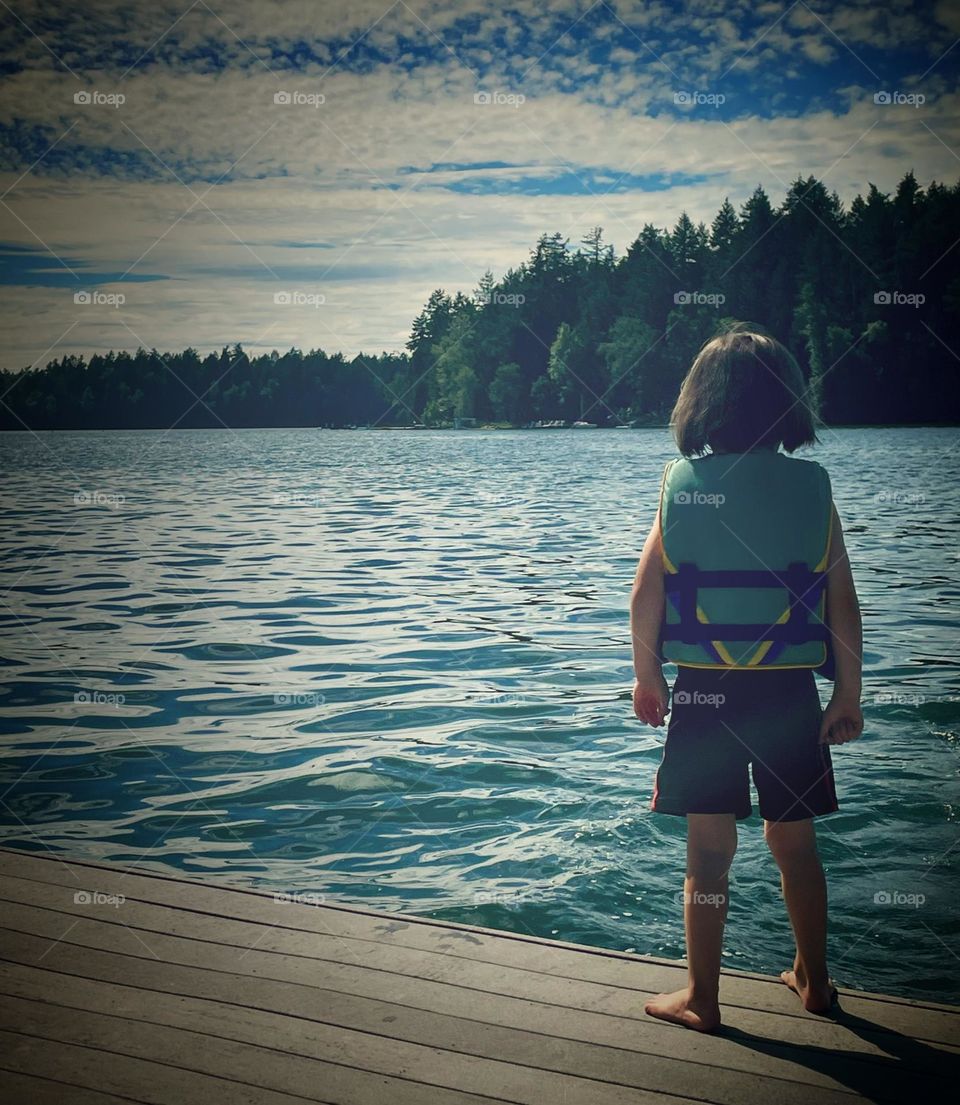 A young child stands on a lakeside dock looking out towards the water in summer