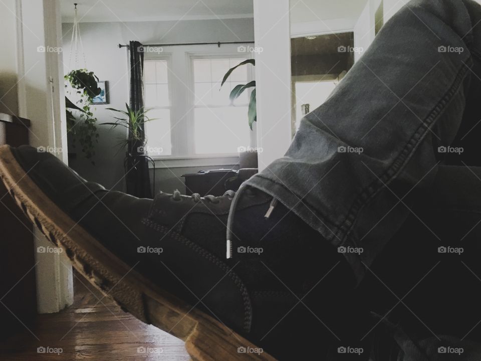 Stylish men’s boot on a man sitting on a sofa in a hardwood-floor room with lush indoor plants and modern interior design 