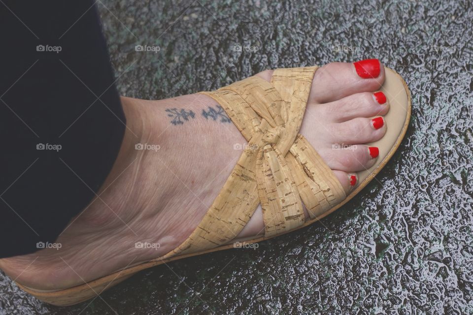 An image of a woman's foot with a tattoo and toes with red nail polish as she stands on the the street.