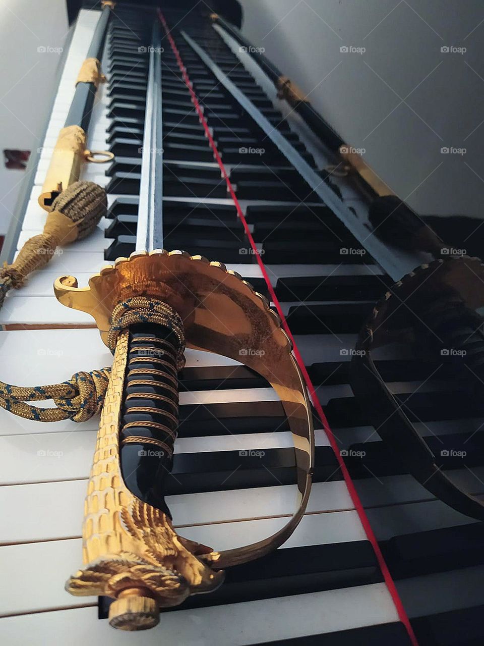 On the wooden keys of the piano lie a metal sword and scabbard.  The sword consists of a metal handle with a bird's head and a metal blade.  The sword reflects off the black wooden surface of the piano.  Ceremonial sword of Italian military aviatio