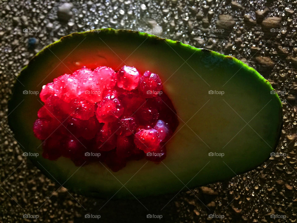 Half a wet green avocado with pomegranate seeds in the hollowed-out center. The avocado is sittting on a wet wood table and is side lit to highlight the drops on the table and show the translucency of the pomegranate seeds. 