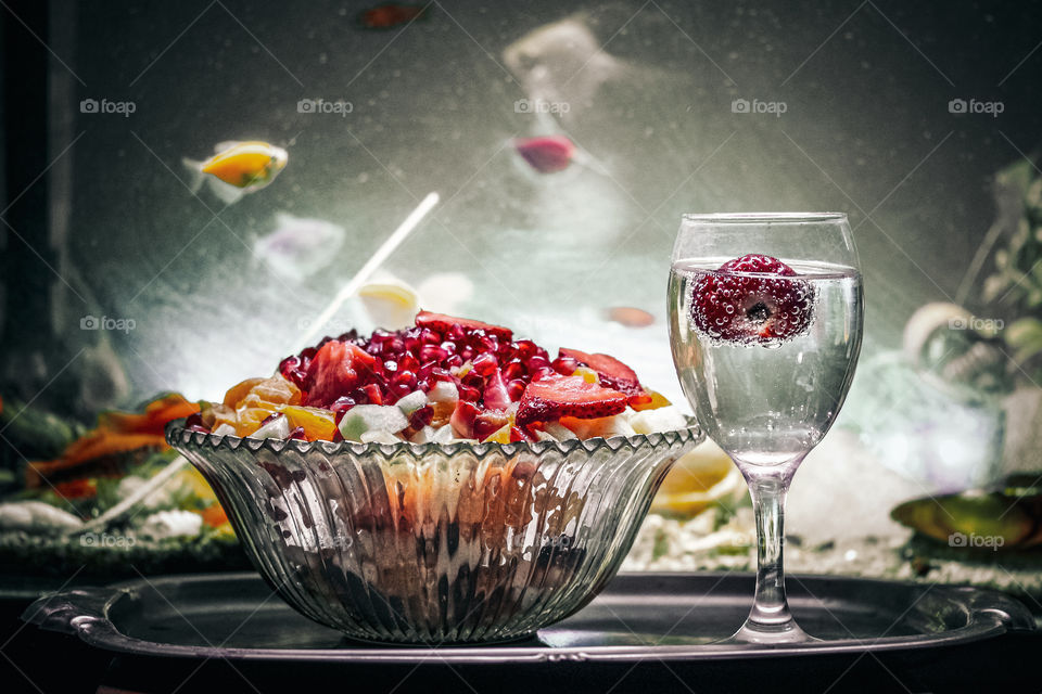 fruit salad and mineral water with a strawberry at the glass of water and aquarium fish swimming in the background of photo