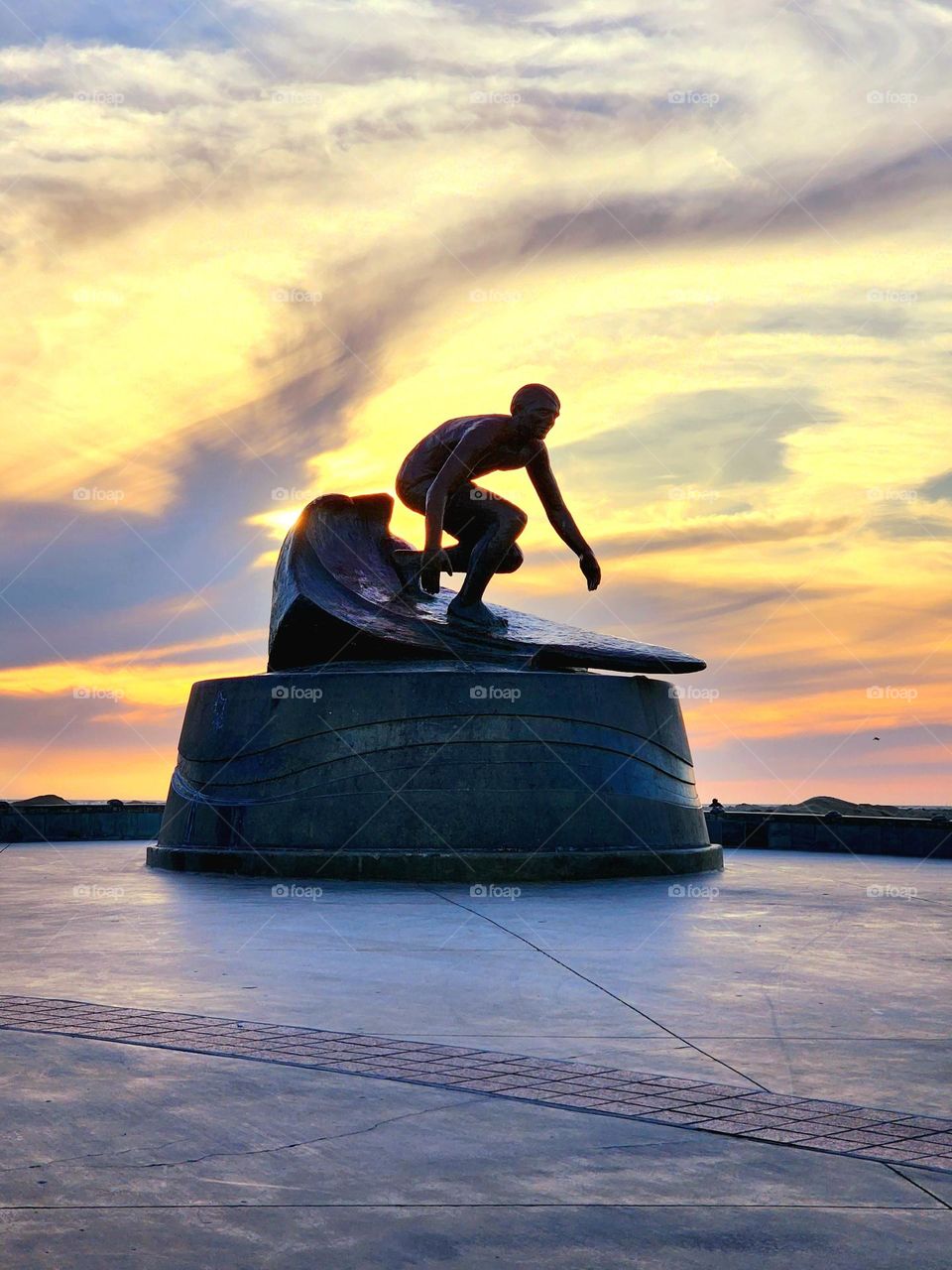 A cloudy sunset over the Pacific Ocean backlights a statue celebrating the great surfers of the area