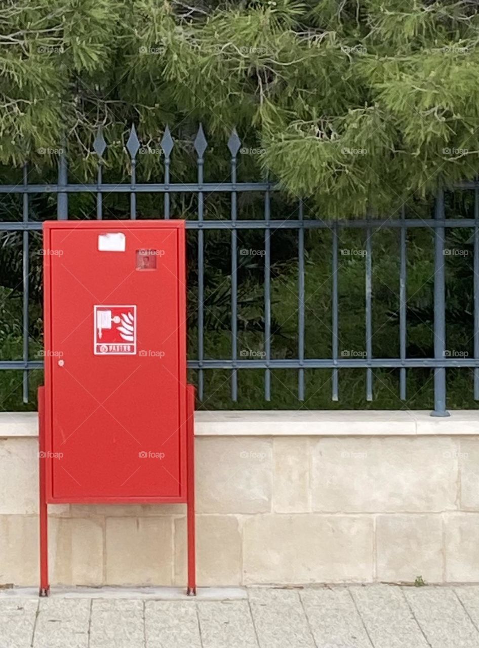 A safety red fire extinguisher box against a dark green fence and green trees.