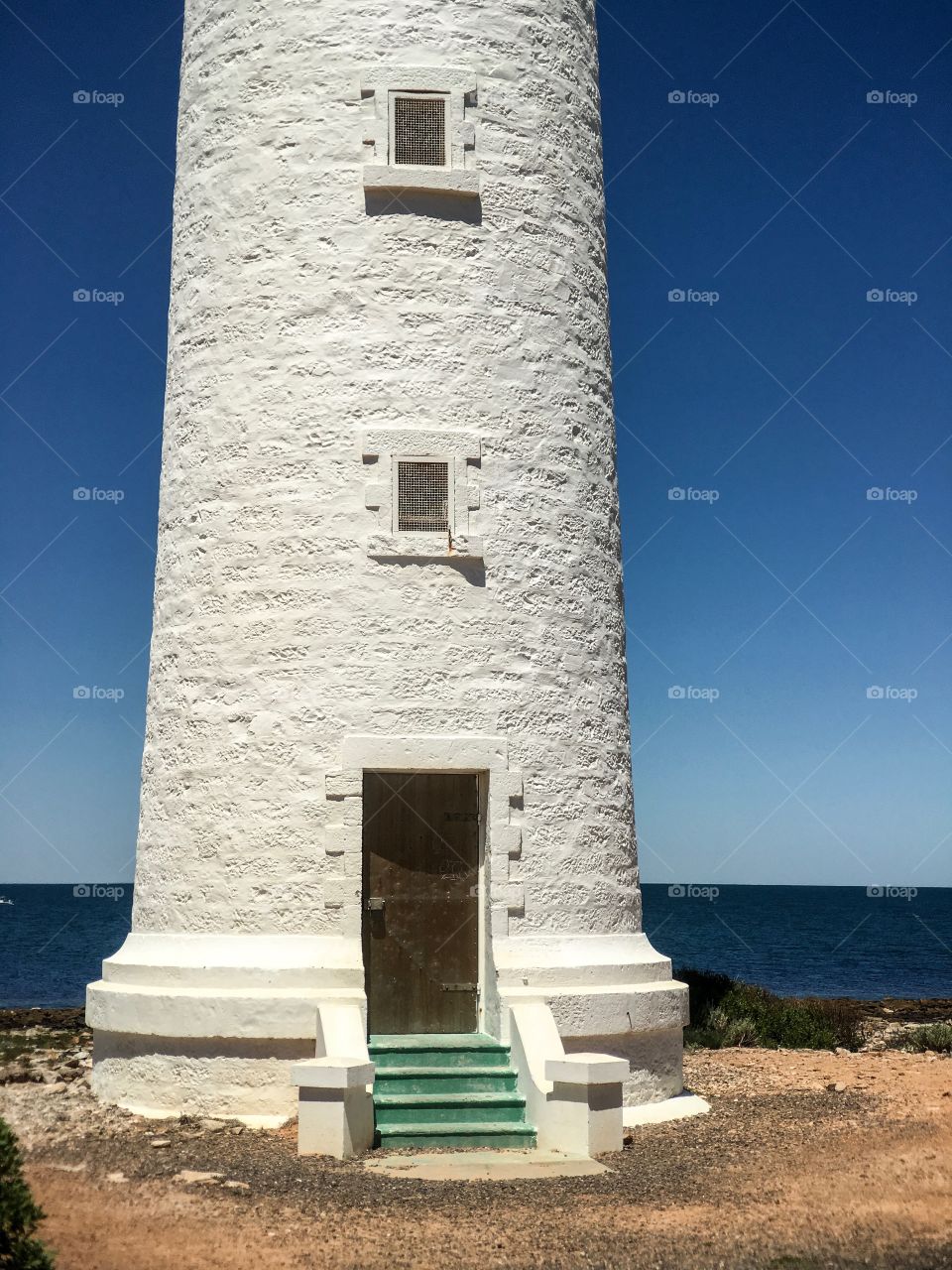 Base of old stone lighthouse, its green stairs and brass door against vivid indigo blue sky and ocean backdrop