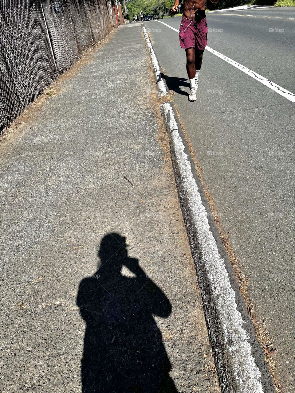 Shadow of photographer/spectator watching man in maroon shorts who is wearing headphones as he runs up a long hill in a bike lane