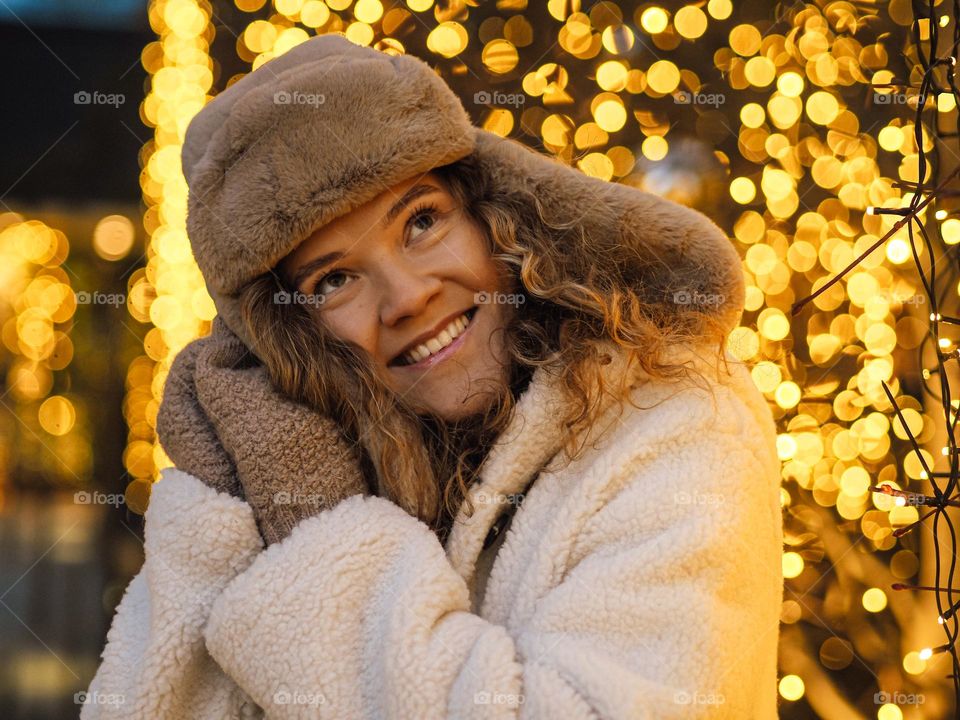 Young beautiful woman with curly hair in fur coat and hat with ear flaps smiling with lights on a background