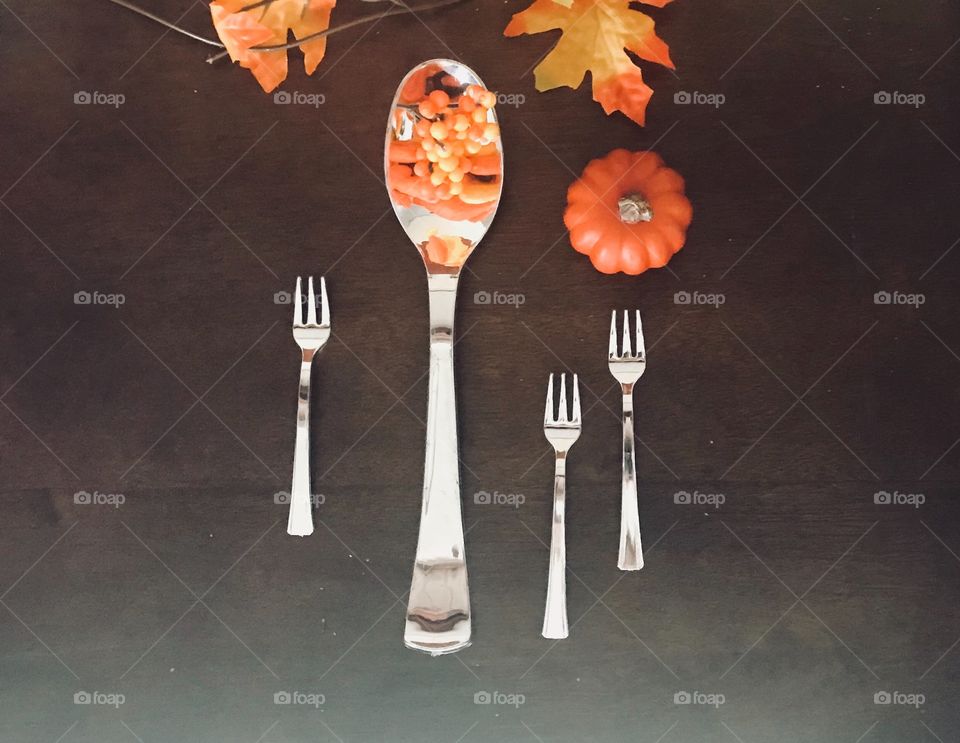 A beautiful table top display for the fall holiday season with a large silver spoon filled with holiday autumn berries surrounded by mini silver forks a mini orange pumpkin and fall colorful garland - orange, yellow, gold, red, brown and green