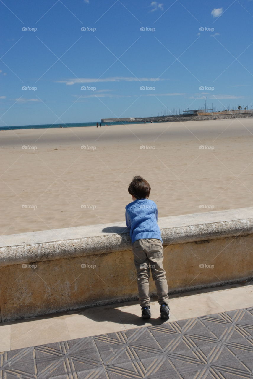 El niño mirando al mar azul al fondo y la arena blanca en un día soleado de verano .