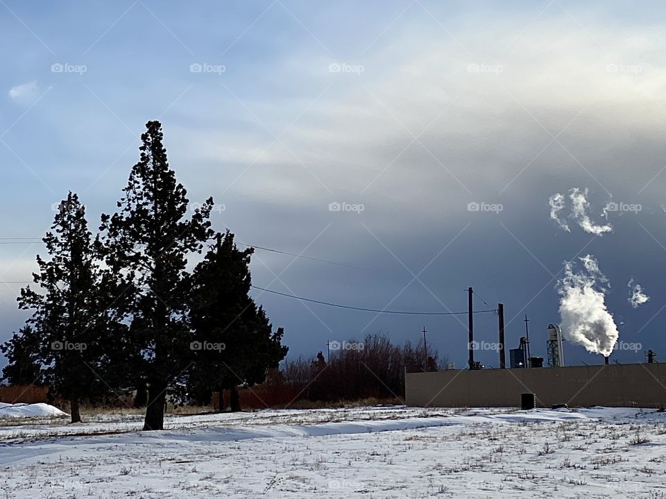 Bright white steam from an industrial plant at the edge of a snow covered field contrasts against a dark and stormy sky. 