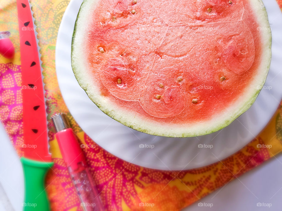 Half of a personal size watermelon on a white plate, on top of a colorful tropical summer place mat, with a watermelon cutting knife and watermelon juice spigot to the left.