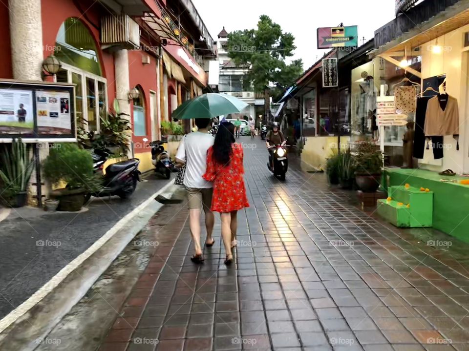 Walking in stride 4. Adult couple with open umbrella. Brick paved street. Shops. Motorcycle. Ubud, BalI. 2018.