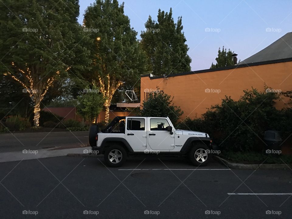 Wide shot of a white four-door Jeep Wrangler against the building and a street that has trees with Christmas lights