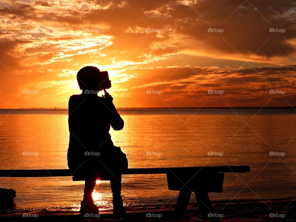 A silhouette of a woman sitting on a picnic table on the beach taking sunset photos 