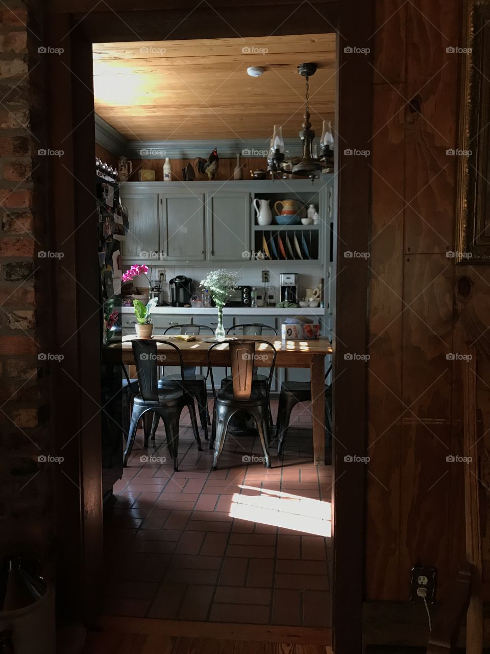 A kitchen. Cozy and homey, brick floors and grey wooden cabinets, flowers on the table, and light pouring through the open window. 