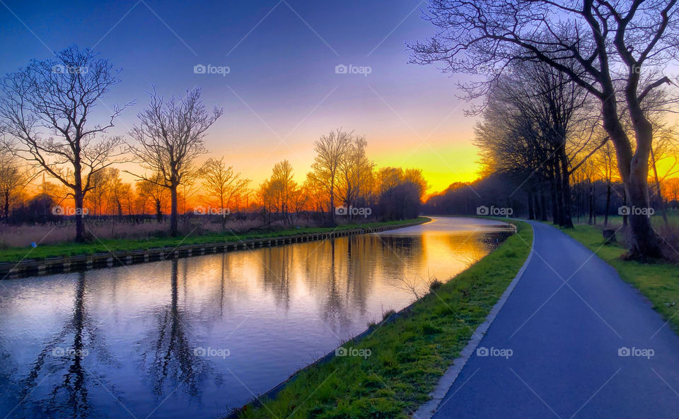 Small road next to a river or canal in a rural Countryside landscape under a colorful and dramatic sunset or sunrise sky