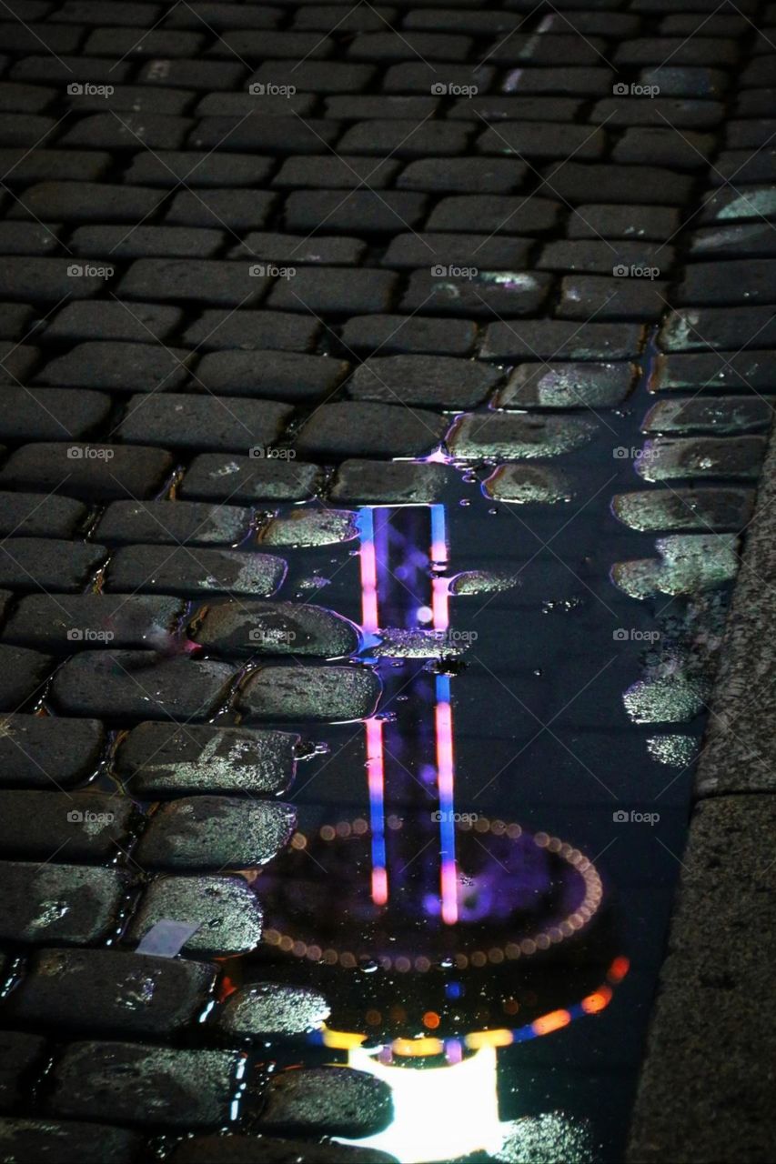 A colorful carousel is reflected in a puddle on cobblestones at night