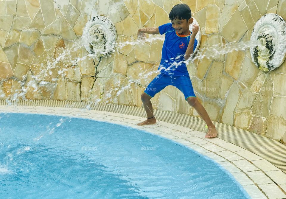 A boy enjoying a lot at swimming pool by doing graceful dance in between two water fountains .