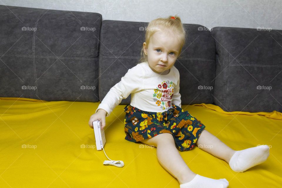 A small, red-haired girl is laughing at home on a gray sofa with purple pillows and a yellow background