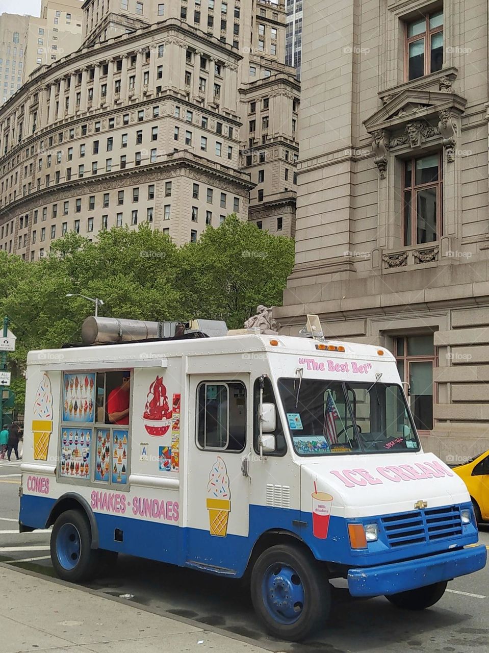 An ice cream parlor waits for customers on the streets of New York. His brightly colored truck contrasts with the color of the surrounding buildings.