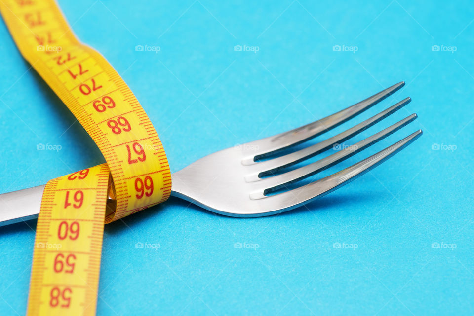 Tape measure around a fork as concept for diet. Fork are wrapped in yellow measuring tape on blue background