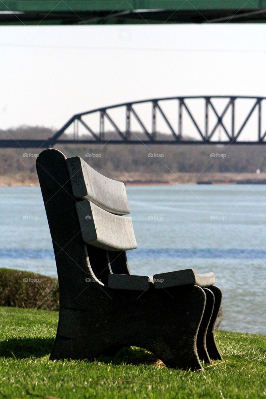 This is a picture of a bench at a park by the Ohio River with a bridge in the background.