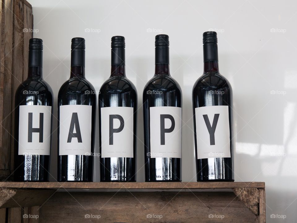 Five black winebottles on a wooden shelf with the word happy written on white labels