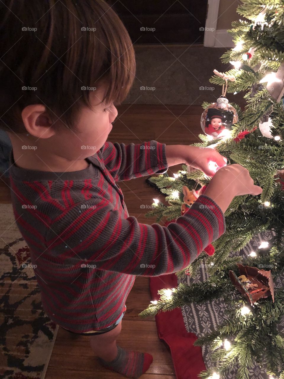 Small boy in his pajamas putting an ornament on the Christmas tree .