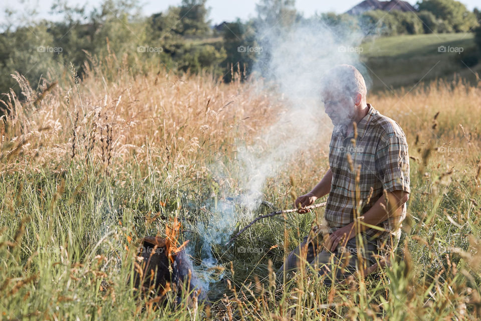 Man starting a campfire, blowing on a fire. Little girl sitting in a grass beside a campfire. Candid people, real moments, authentic situations