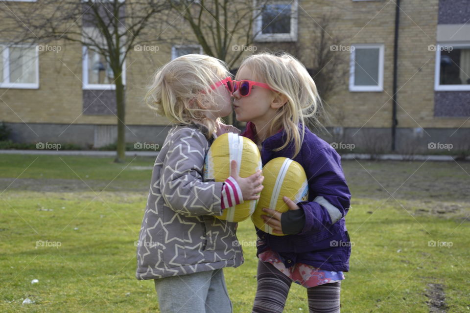 Two little blonde girls with toy kissing in front of building