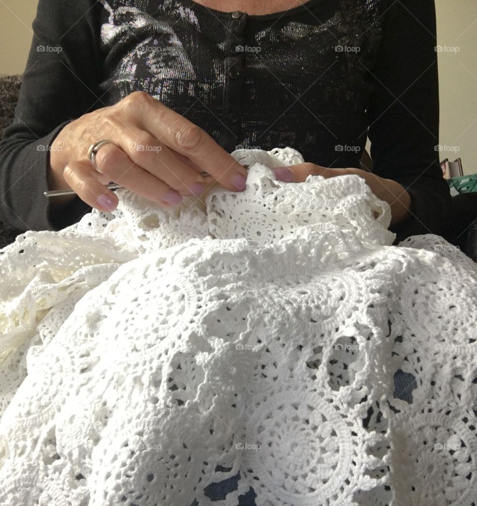 Woman sitting crocheting fine lace tablecloth using fine white crochet cotton thread and steel crochet hook, needlework, work is on her lap